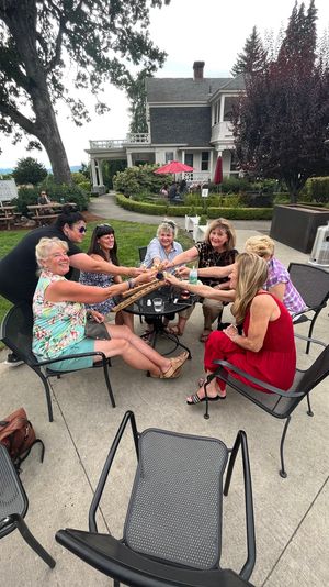 Group of women toasting with small glasses around a metal patio table on a landscaped lawn in front of a Victorian-style house