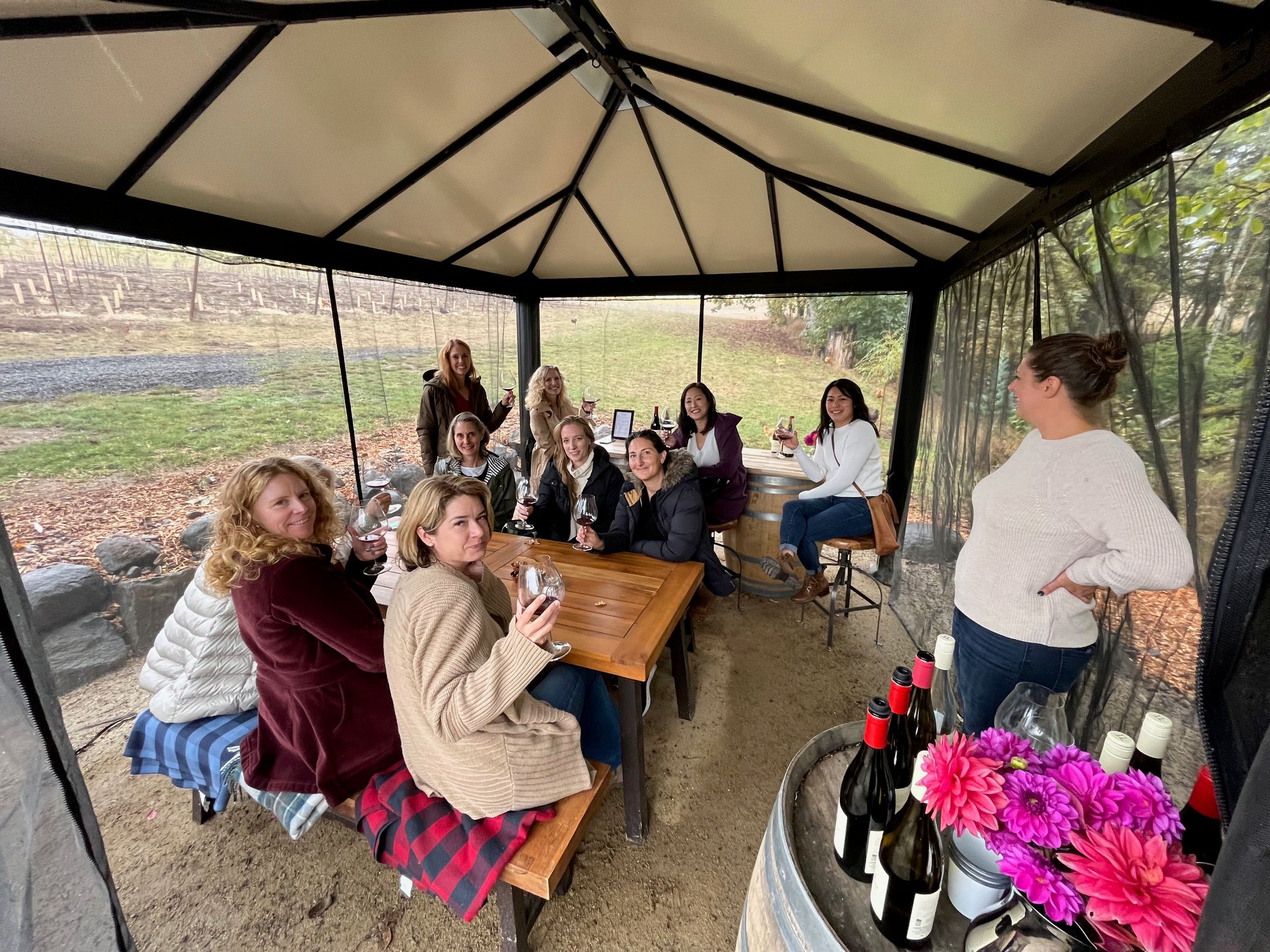 Cheerful group enjoying an outdoor wine tasting in a covered vineyard gazebo, seated at a wooden table with wine glasses and bottles on a barrel, autumn vines visible outside.