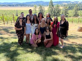 Cheerful group of friends posing for a sunlit summer photo in a vineyard, with rows of grapevines, trees and rolling hills under a clear blue sky.