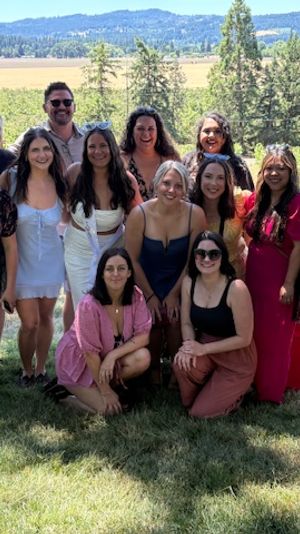 Cheerful group of friends posing for a sunlit summer photo in a vineyard, with rows of grapevines, trees and rolling hills under a clear blue sky.