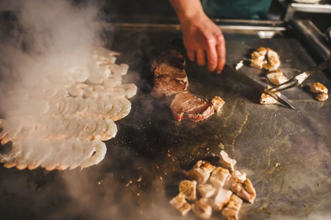 Sizzling teppanyaki: a cook’s hand sears steak medallions and a row of shrimp on a steaming flat-top griddle in a restaurant kitchen.