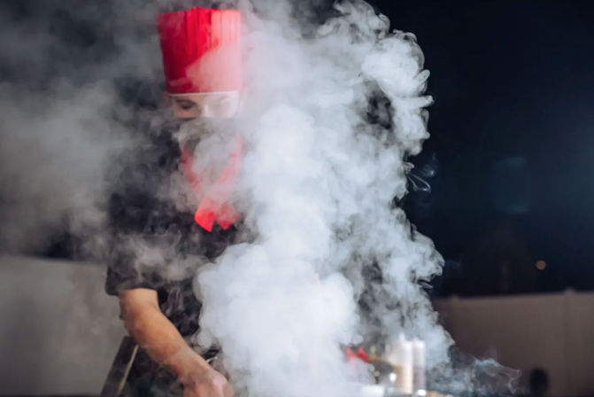 Smoky nighttime outdoor grill scene with a chef in a red toque and matching neckerchief, hands working over a sizzling grate as thick white cooking smoke billows up.