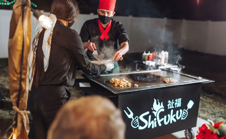 Masked chef in a red hat cooks sizzling teppanyaki on a flat-top grill at an outdoor night market, steam rising as he hands a plate to a waiting customer.