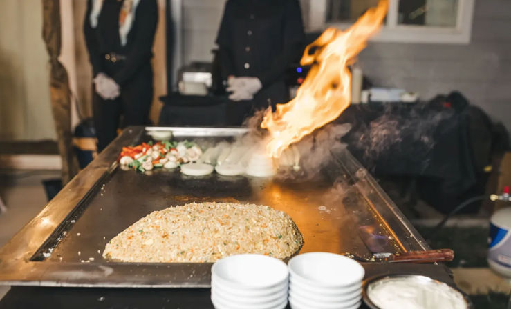 Outdoor teppanyaki-style flat-top griddle at a catering event, mound of fried rice with sliced onions and vegetables and a dramatic flame sizzling on the hibachi grill