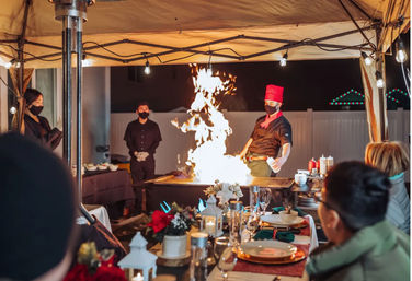 Masked chef in red hat flambéing on a teppanyaki grill under a lit outdoor tent while masked servers and guests watch a festive, candlelit backyard dinner at night