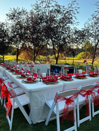 Outdoor garden reception at sunset — long white-clothed banquet table on green lawn set with gold chargers, red napkins, glassware and candles; white folding chairs tied with red bows, trees in background.