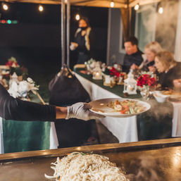 Gloved server passes a plate of vegetables and shrimp over a steaming flat-top grill piled with noodles at an outdoor, candlelit catered dinner under string lights.