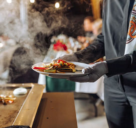 Gloved server holding a steaming plate of grilled vegetables at a catered event, blurred banquet tables and warm string lights in the background.