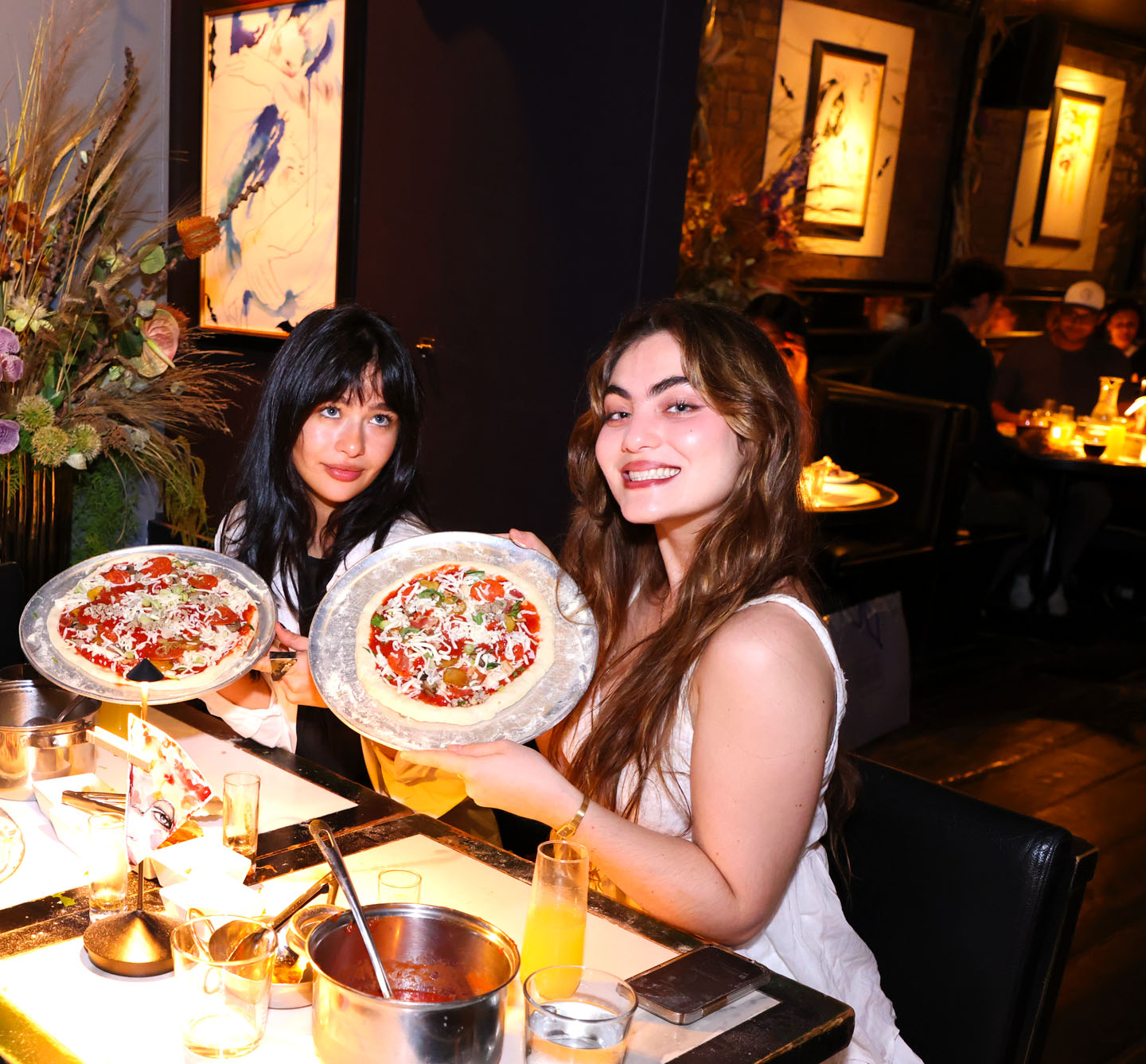 Two smiling women at a cozy, dimly lit restaurant table holding personal pizzas on metal trays, with a sauce pot, drinks and tableware on the table and artwork on the walls in the background.
