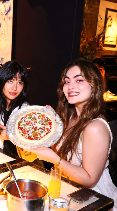 Two smiling women at a cozy, dimly lit restaurant table holding personal pizzas on metal trays, with a sauce pot, drinks and tableware on the table and artwork on the walls in the background.