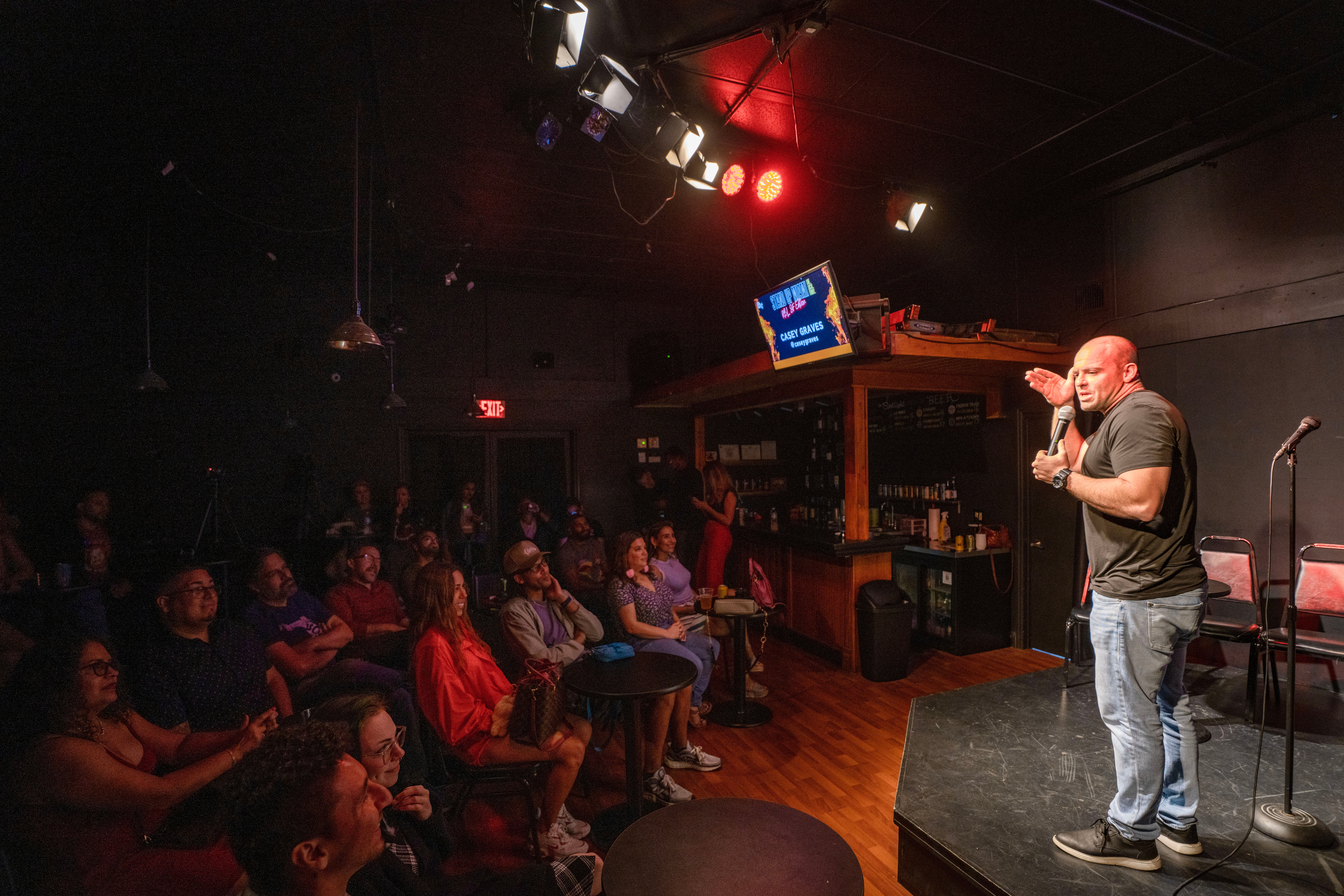 Live stand-up comedy in an intimate comedy club — comedian on stage with a microphone, laughing audience at small tables, bar and red stage lights in the background.