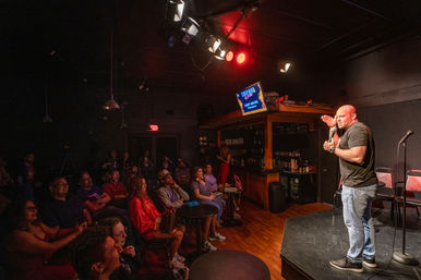 Live stand-up comedy in an intimate comedy club — comedian on stage with a microphone, laughing audience at small tables, bar and red stage lights in the background.