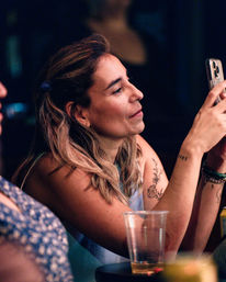 Woman with floral arm tattoo checking her smartphone at a dimly lit bar table with a plastic cup and drinks nearby — casual nightlife scene.