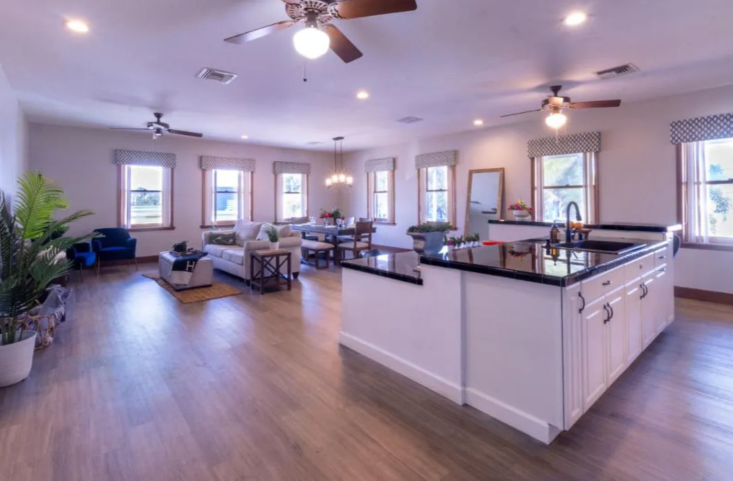 Bright open-concept living room and kitchen with a large white island and black countertops, hardwood floors, ceiling fans, dining table, cozy sofa seating, potted plants, and sunlit windows.