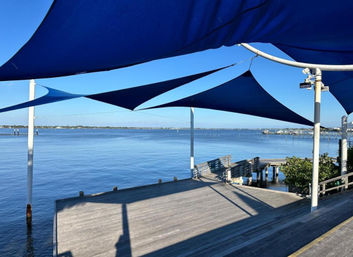 Sunlit wooden waterfront pier with blue triangular shade sails overhead, benches and a small dock extending into a calm blue bay under a clear sky