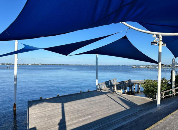 Sunlit wooden waterfront pier with blue triangular shade sails overhead, benches and a small dock extending into a calm blue bay under a clear sky