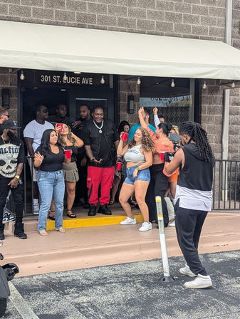 Outdoor party on St. Lucie Ave — a lively group holding red cups dancing and posing outside a storefront while a videographer films.