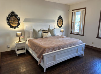 Bright cozy bedroom with a white wooden king bed dressed in a blush quilt and decorative pillows, matching nightstands with lamps, dark hardwood floors and two ornate wall plaques near a window.
