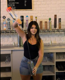 Bartender performing a dramatic long pour into a cocktail shaker in a white-brick bar with a row of beer taps.