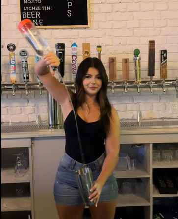 Bartender performing a dramatic long pour into a cocktail shaker in a white-brick bar with a row of beer taps.