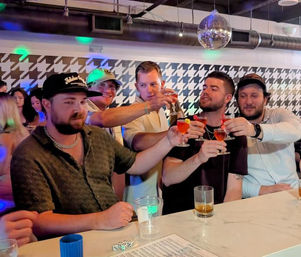 Group of friends toasting with pink cocktails at a lively bar counter under a disco ball, houndstooth-pattern wall and drinks on a marble bar top — upbeat nightlife scene.