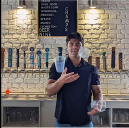 Smiling bartender juggling clear plastic cups in a craft beer taproom, in front of a white brick wall with a row of beer taps and a chalkboard menu listing cocktails and beer.