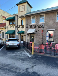 Downtown storefront with small clock tower and green awnings, white SUV parked by a door labeled “Private Entrance,” fenced patio with pink cafe chairs.
