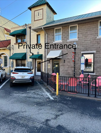 Downtown storefront with small clock tower and green awnings, white SUV parked by a door labeled “Private Entrance,” fenced patio with pink cafe chairs.