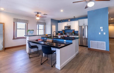 Bright modern open-plan kitchen with blue accent wall, black granite island breakfast bar, two black chairs, stainless steel appliances and wood-look floors