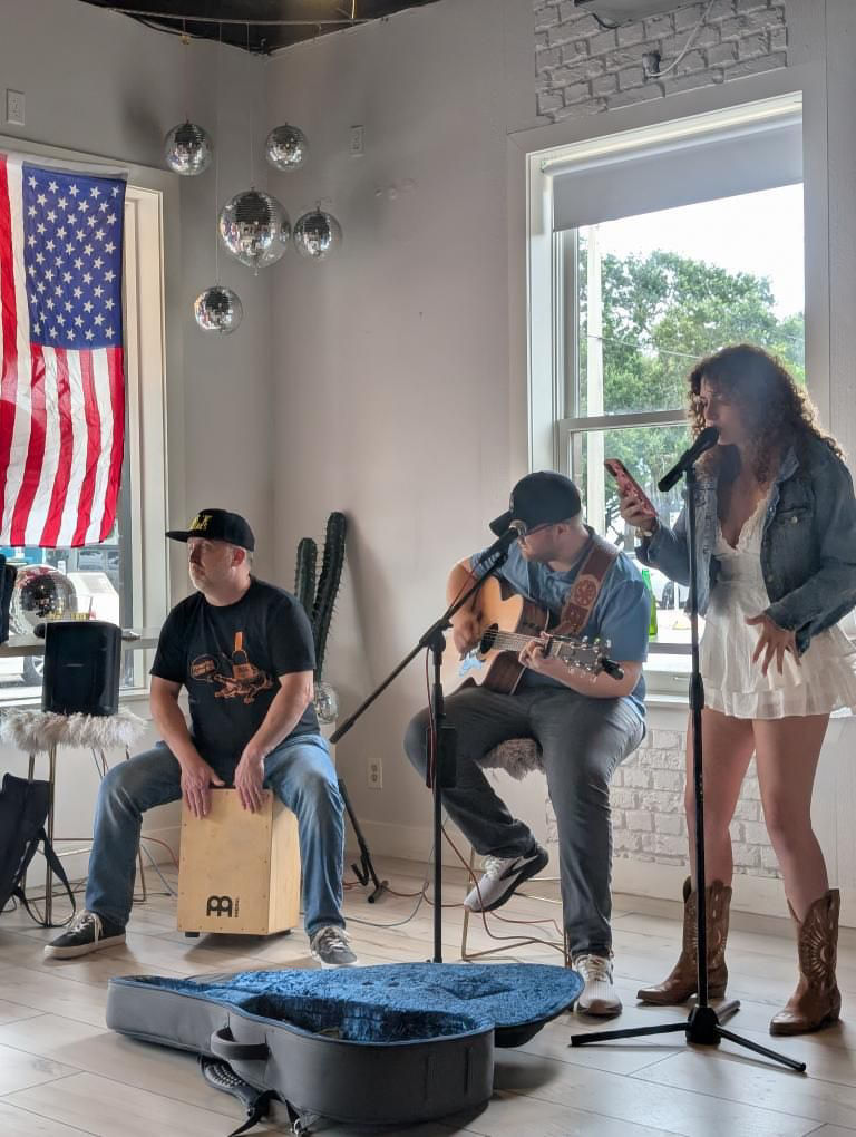 Three-person acoustic band performing an indoor live music set at a bright local cafe — female singer in white dress and cowboy boots at a microphone, seated guitarist on stool, percussionist playing a cajon, open guitar case on floor, American flag and hanging disco balls in the windowed background.
