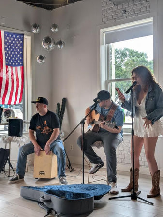 Three-person acoustic band performing an indoor live music set at a bright local cafe — female singer in white dress and cowboy boots at a microphone, seated guitarist on stool, percussionist playing a cajon, open guitar case on floor, American flag and hanging disco balls in the windowed background.