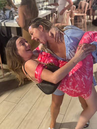 Two women laughing during a lively night out at an indoor bar, one dipping her friend in a bright pink patterned dress.