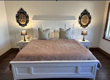 Cozy farmhouse bedroom with large white wooden bed dressed in a dusty-rose quilt, three neutral throw pillows, matching white nightstands with lamps, ornate round framed wall art, hardwood floor and a sunlit window.