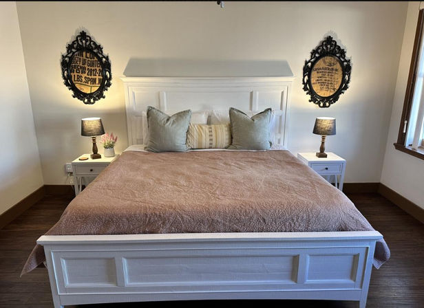 Cozy farmhouse bedroom with large white wooden bed dressed in a dusty-rose quilt, three neutral throw pillows, matching white nightstands with lamps, ornate round framed wall art, hardwood floor and a sunlit window.