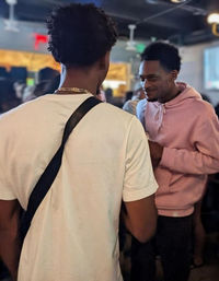 Two young men chatting in a crowded indoor bar with a nightlife vibe — one facing away in a white tee and crossbody strap, the other smiling in a pink hoodie.