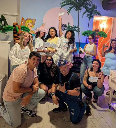 Group photo of a cheerful group at an indoor tropical-themed party with inflatable palm trees, colorful mural backdrop, party props and signs, people smiling and posing on a wood floor.