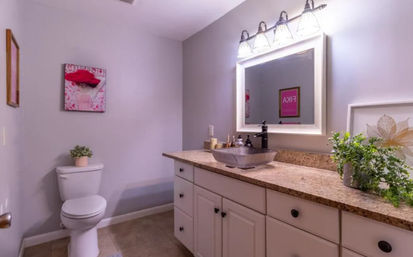 Contemporary residential bathroom with granite countertop and white vanity, glass vessel sink with black faucet, illuminated framed mirror, toilet with potted plant and decorative wall art.