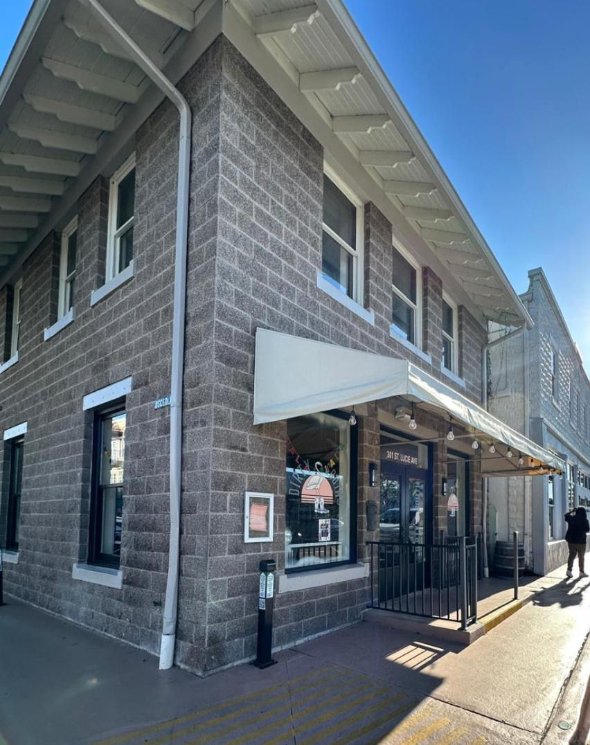 Sunny corner two-story gray cinderblock storefront with a white awning, string lights and sidewalk entrance on a downtown street, a person walking past