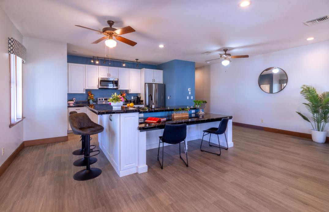 Bright open-concept modern kitchen interior with blue accent wall, white cabinets, stainless steel appliances, black granite island with bar stools, ceiling fans, wood-look flooring, round wall mirror and potted plant.