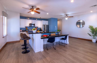 Bright open-concept modern kitchen interior with blue accent wall, white cabinets, stainless steel appliances, black granite island with bar stools, ceiling fans, wood-look flooring, round wall mirror and potted plant.