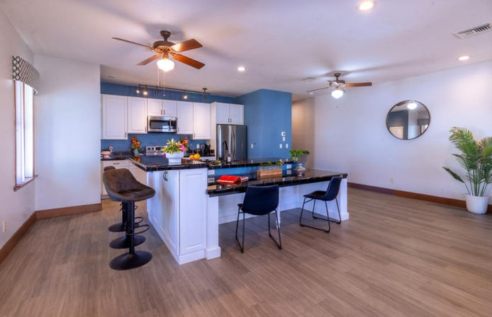 Bright open-concept modern kitchen interior with blue accent wall, white cabinets, stainless steel appliances, black granite island with bar stools, ceiling fans, wood-look flooring, round wall mirror and potted plant.
