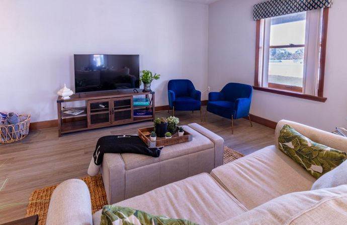 Bright living room with beige sectional and ottoman topped by a wooden tray of succulents, two blue velvet accent chairs, wooden media console with flat-screen TV, woven rug, and a large window showing a water view.
