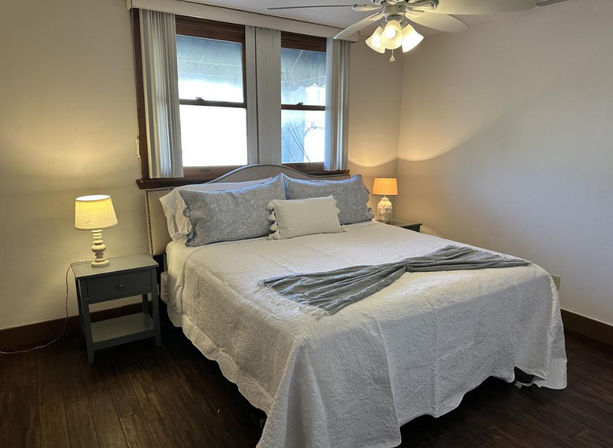 Calming cozy bedroom interior with a large white quilted bed, blue patterned pillows, gray throw, two bedside tables with lamps, hardwood floors, ceiling fan and double window with vertical blinds.
