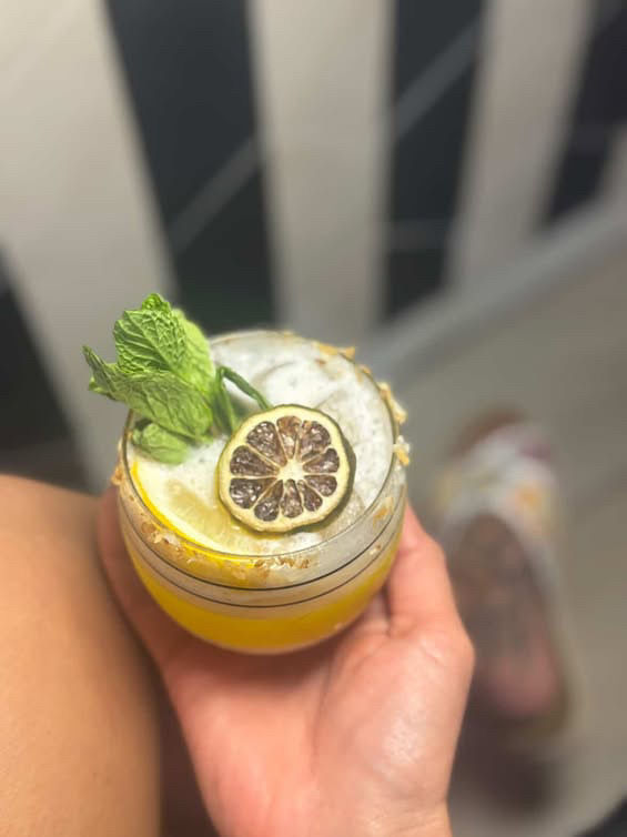 Bright yellow citrus cocktail in a short glass held in hand, topped with crushed ice, a mint sprig and dried lemon wheel on a sugared rim, with a blurred striped background