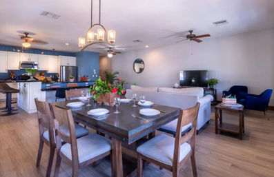 Bright open-plan living space with rustic wooden dining table set for four and floral centerpiece, kitchen island with bar stools, white cabinets and stainless appliances, beige sofa, TV and blue accent chairs