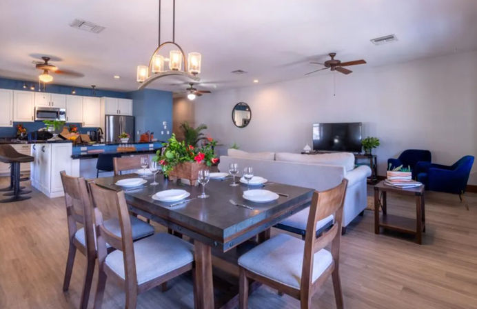 Bright open-plan living space with rustic wooden dining table set for four and floral centerpiece, kitchen island with bar stools, white cabinets and stainless appliances, beige sofa, TV and blue accent chairs