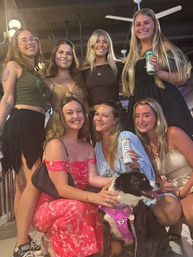 Seven smiling young women posing on a covered patio at night, wearing summer dresses and casual tops, several holding canned drinks while one pets a black-and-white dog in a purple harness.