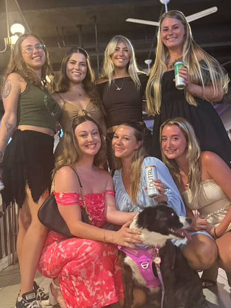 Seven smiling young women posing on a covered patio at night, wearing summer dresses and casual tops, several holding canned drinks while one pets a black-and-white dog in a purple harness.