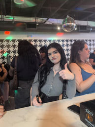 Playful young woman giving a thumbs-up at a marble bar counter in a lively indoor lounge with a disco ball, houndstooth wall pattern and other patrons.