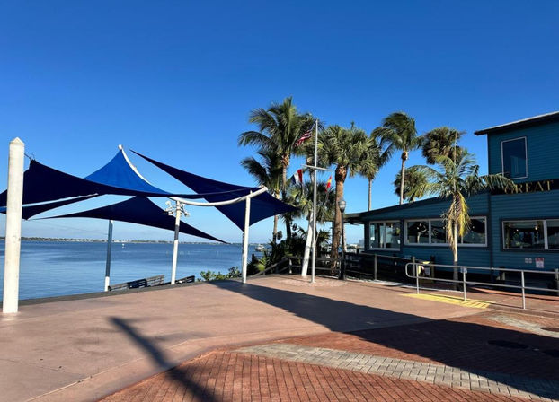 Sun-drenched waterfront boardwalk with blue triangular shade sails, swaying palm trees and a turquoise seaside restaurant overlooking a calm bay under a clear blue sky.